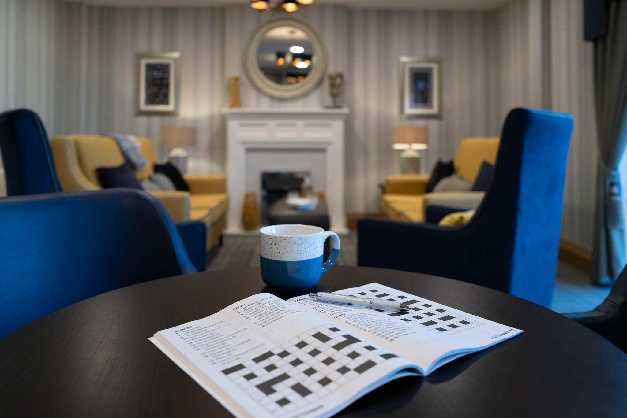 Crossword puzzle on a black table with blue mug. Blurred background of a lounge with fireplace, circle mirror, mustard sofas and navy blue armchairs.