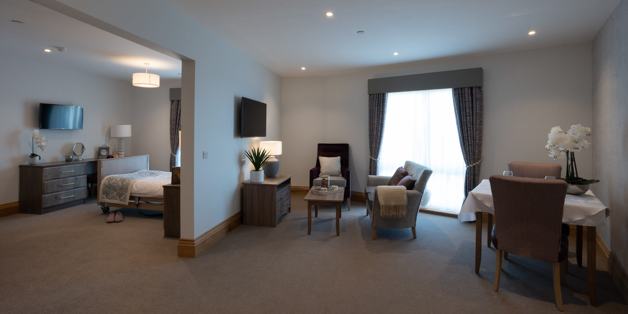 A bedroom suite with archway to lounge. Grey sofa and black armchair with coffee table. Dressed dining room table and chairs. Two sets of patterned curtains and matching bed runner. Vanity table with TV above.