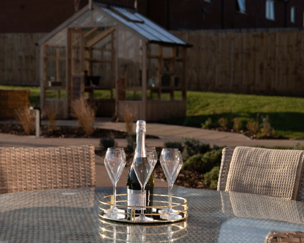 A garden table with a green house in the background. Four glasses and a bottle of Champagne.