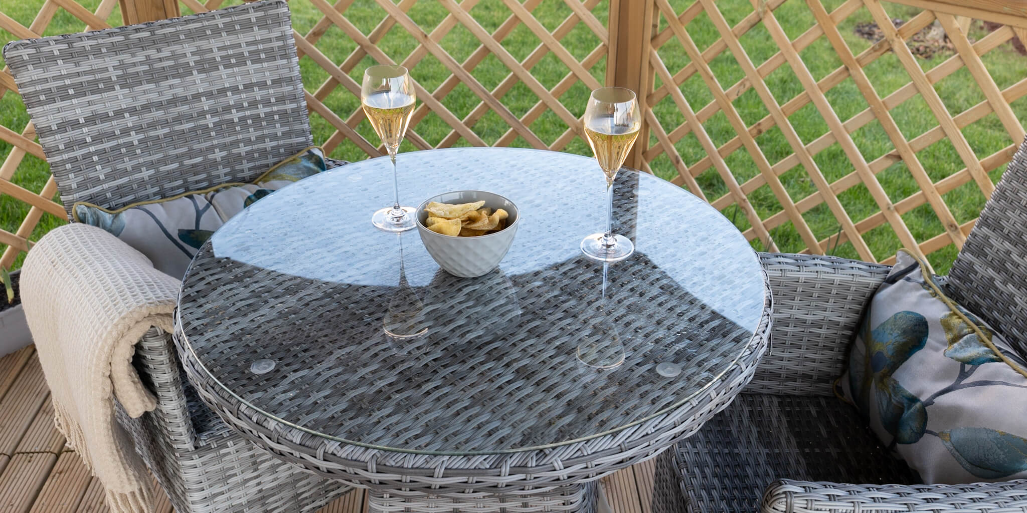 Garden table and chairs with floral pillows and brown/cream blanket. Bowl of crisps and two wine glasses.