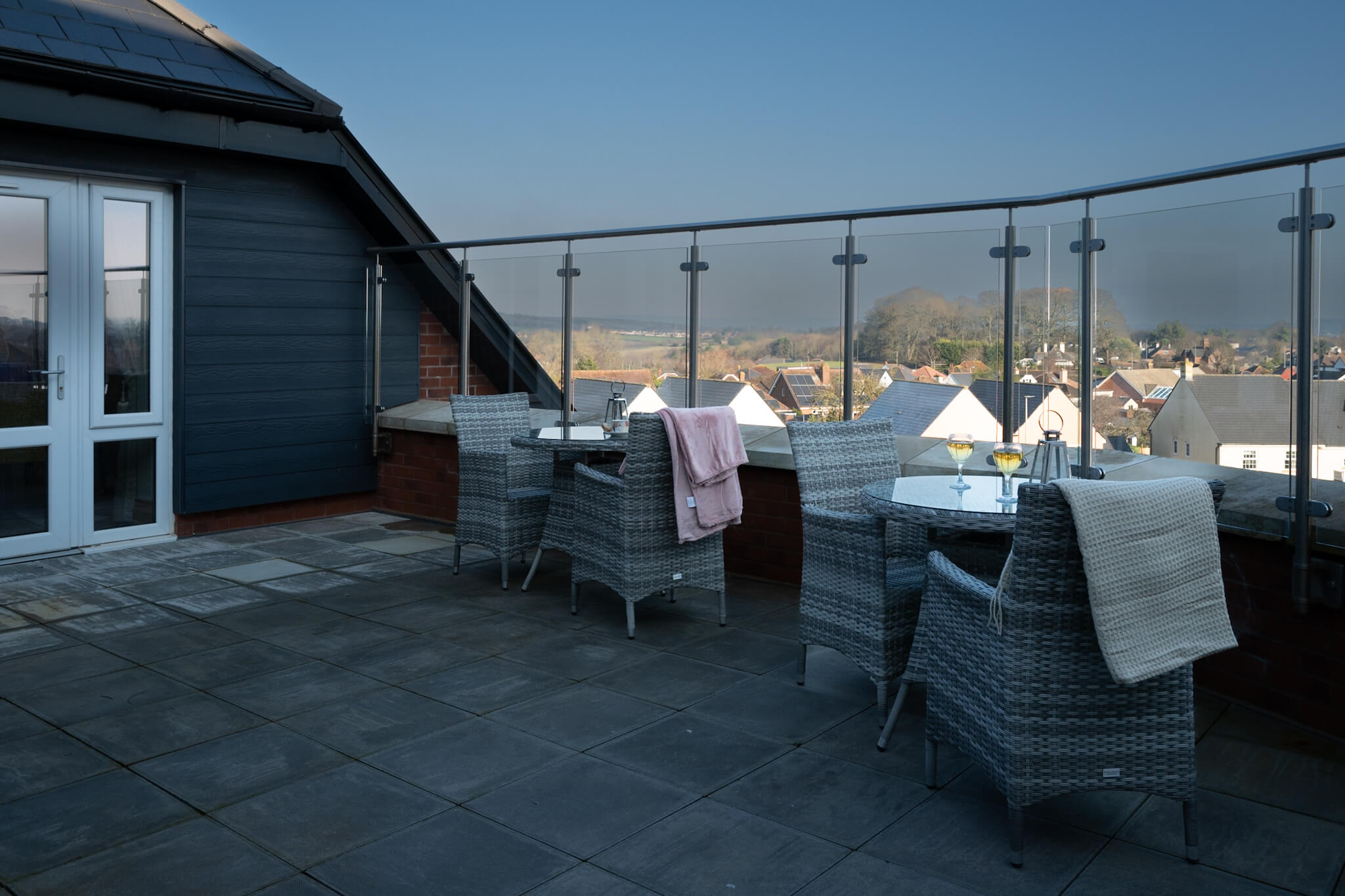 Roof terrace with two sets of garden dining tables. Blankets on back of chairs. Glass fencing. Two wine glasses.