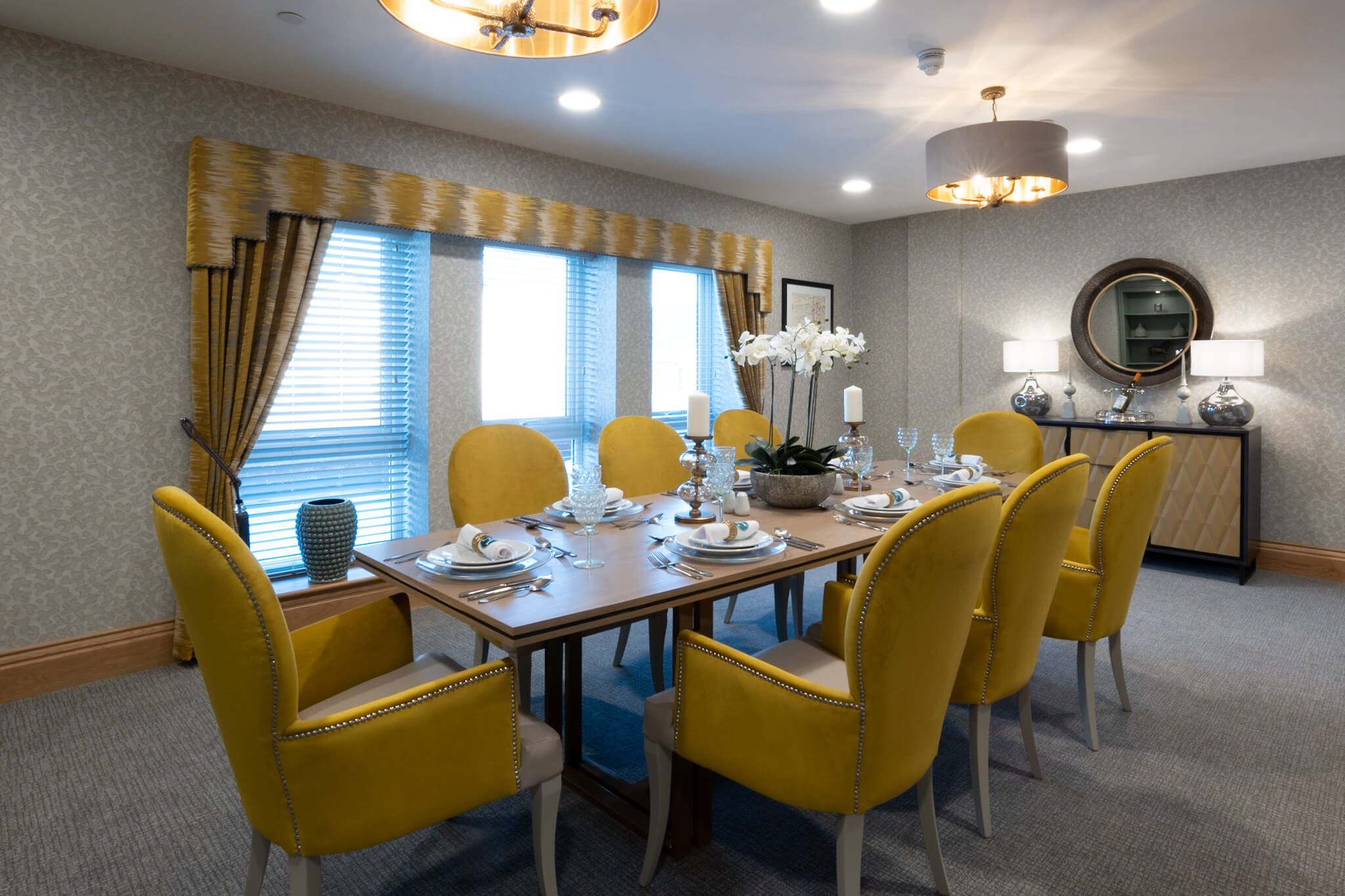 Dressed table for eight people. Yellow dining room chairs with matching curtains. Console table with circle mirror above.
