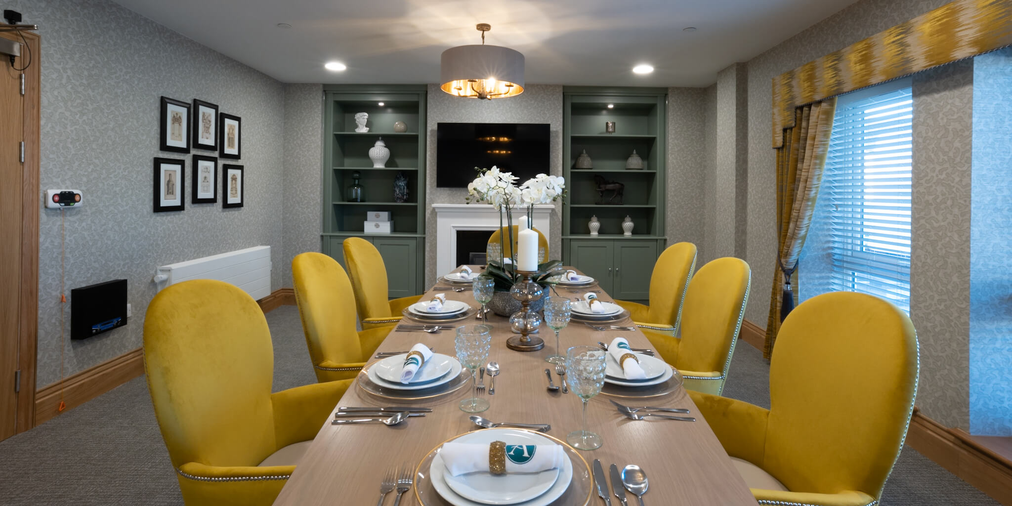 Private dining room. Dressed table with yellow dining chairs. Matching yellow curtains. Flat screen TV on wall with sage green bookcases on wither side. Six black framed photos on the wall.
