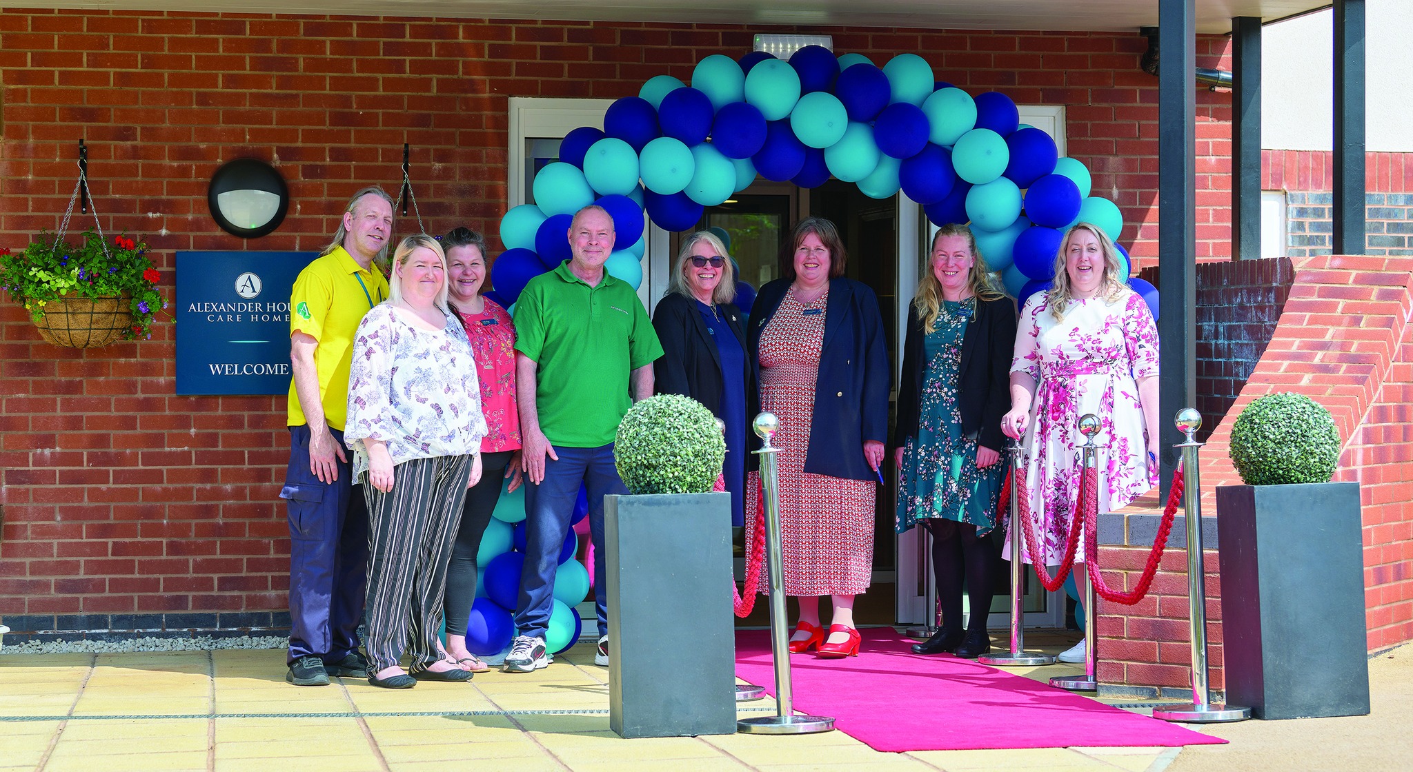 Six females and two males smiling in front of a light blue and dark blue balloon arch with a red carpet.