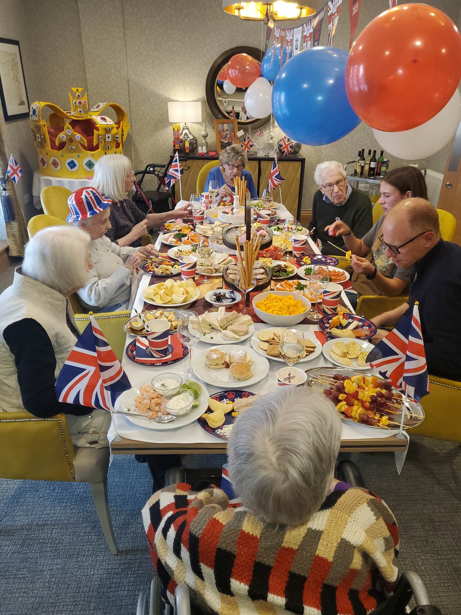 A group of residents and loved ones around a table eating a selection of food. Balloons and Union Jack flags.