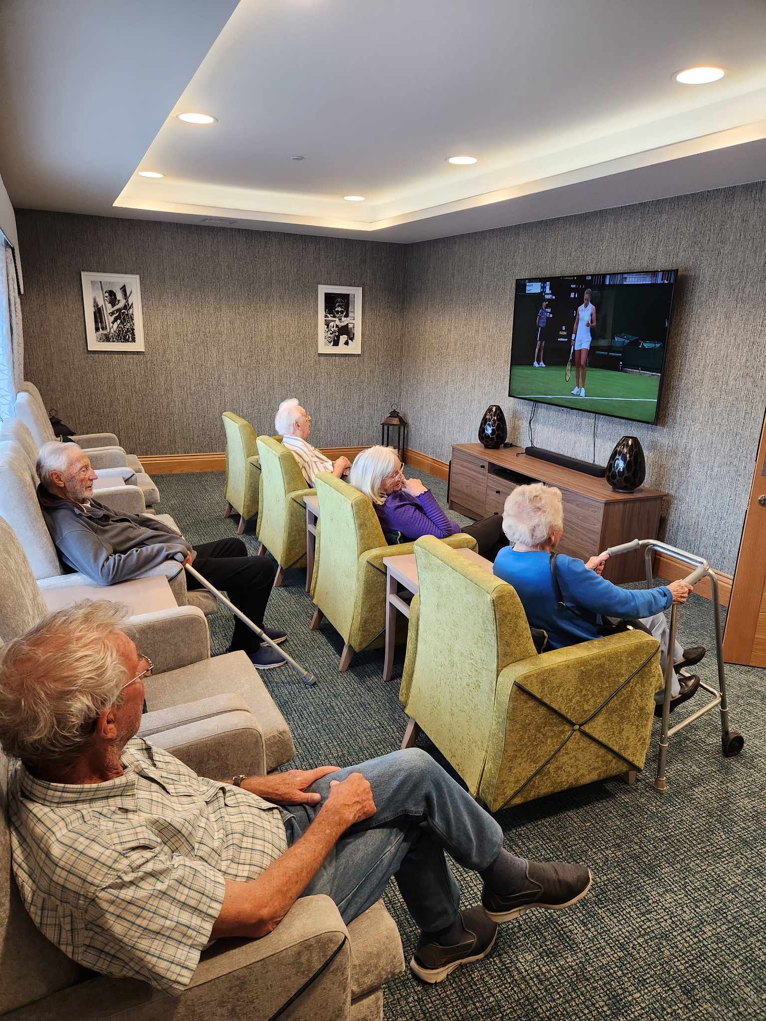 Two male residents in the back row armchairs. Three female residents in the front row green armchairs watching the tennis on the flatscreen TV.